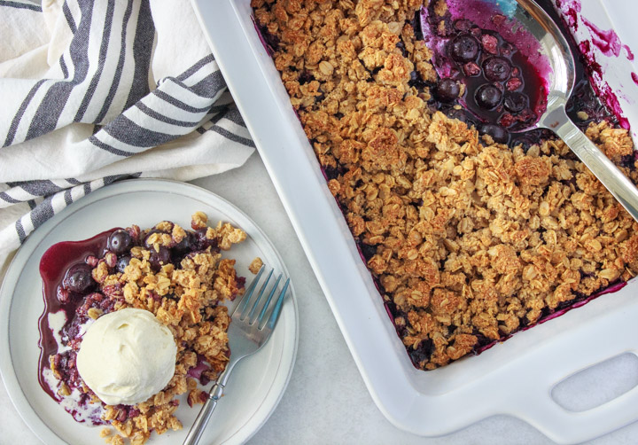 A dish of blueberry crisp with a little dessert plate topped with blueberry crisp served and ice cream.
